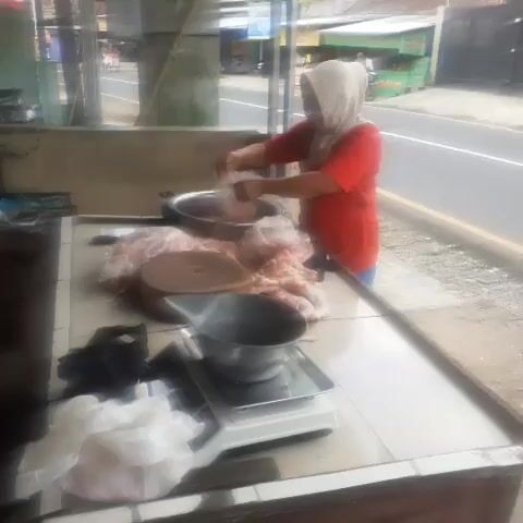 A person wearing a headscarf and a red shirt is standing at a butcher's stall. They are preparing meat, which is spread out on a table. There is a scale on the counter, and various utensils and containers are visible. The stall is situated next to a road, with buildings and vegetation in the background. The video appears to be an excerpt from a longer recording, as indicated by the time stamp.