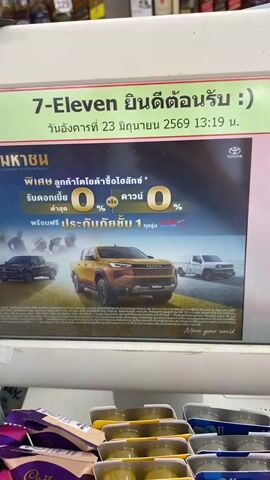 The video shows a close-up of a 7-Eleven receipt displayed on a screen or sign. The receipt indicates a greeting and provides a date and time: Tuesday, June 23, 2569 at 13:19. Below this, an advertisement for Toyota vehicles is visible, featuring several trucks and offering promotions like 0% down payment and 0% interest for a certain period, along with free insurance. A hand with red nail polish points to the receipt and advertisement. The background shows shelves stocked with bottles, likely in a convenience store setting. The timestamp progresses slightly throughout the short clip.