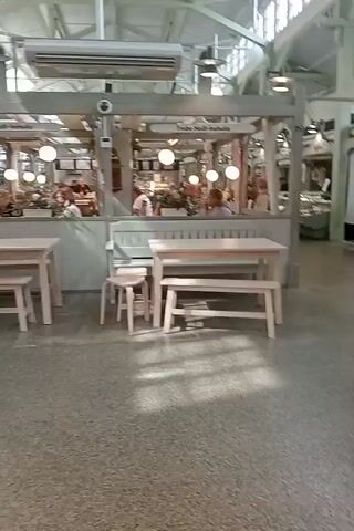 The video shows an indoor market with stalls and seating areas. In the foreground, there is a light-colored wooden table with two benches, and to the left, two wooden stools. The stall behind the table has a glass display case with various items inside and is illuminated by warm pendant lights. To the left of this stall, another stall is visible with more display cases and items. Further in the background, there are several more stalls with similar display cases and architectural features such as arched windows and ceiling beams. The floor is a mottled brown material. The lighting is bright and overhead. The camera is moving, suggesting someone is walking through the market. No specific actions or interactions are clearly discernible, though the presence of stalls suggests commerce.