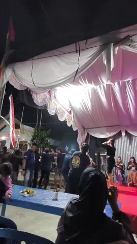 The video shows an outdoor evening event under a large white tent. A group of people are gathered, some standing and watching a performance on a stage. The stage is covered in a red carpet and lit by bright lights. One performer, dressed in black, is on stage, gesturing with their hands. In the foreground, a person wearing a black hijab is seated at a blue table, holding a phone. Other people are visible in the background, including children and adults mingling. Flags are displayed on poles to the left of the tent. Food items are on the blue table.