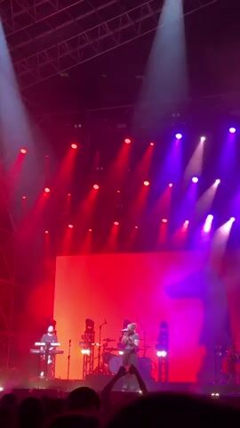 A singer stands on stage, illuminated by red and purple lights, performing for a large crowd. A musician to her left plays a keyboard. There are drums and other equipment behind them. The stage features a large red screen in the background. The video is taken from the crowd, with many hands visible in the foreground.