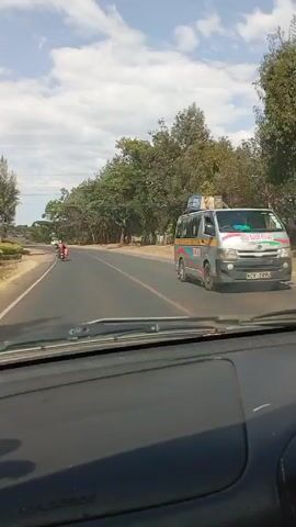The video, shot from the interior of a car driving on a road, shows the roadside. The road is paved and marked with a double yellow line down the center. The roadside is lined with trees and shrubs. There are some houses visible in the background. A motorcycle is traveling in the same direction as the car. Another car is further up the road in front of the motorcycle. A white pickup truck is parked on the side of the road. The sky is blue with white clouds.