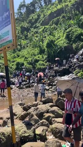 A crowded natural scene with a waterfall cascading down rocks. Many people, appearing to be tourists, are positioned on and around the rocks, some wading in the water, others observing the waterfall. The environment is lush with green vegetation covering the slopes. Umbrellas are visible, suggesting sunny weather. The people are dressed in casual clothing. Some individuals are actively interacting with the water, possibly bathing or cooling off. The overall impression is a popular tourist destination during a busy period.