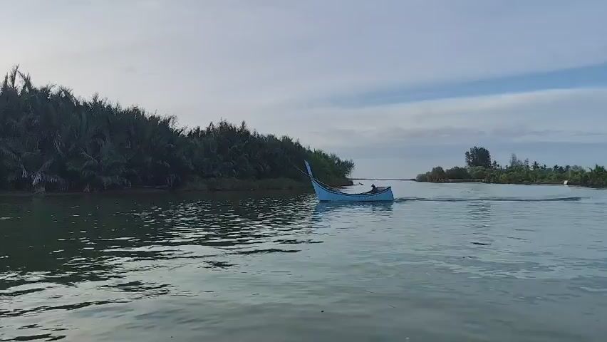 A motorboat travels down a river with a person inside. The boat is blue and appears to be made of wood. The river is surrounded by lush green vegetation on both sides. The sky is overcast with some clouds visible. The water is calm and reflects the sky and trees. The boat is leaving a wake behind it, indicating it is moving.