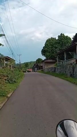 The video shows a first-person perspective of someone riding a motorcycle on a paved road. On the left side of the road, there are houses, some with wooden walls and visible foundations. A grassy area runs alongside the road, with some vegetation. Overhead, power lines and utility poles are visible. On the right side of the road, there are more houses, some with stone foundations and wooden structures, as well as trees and foliage. In the distance, there are more houses, trees, and hills, suggesting a rural or semi-rural environment. The sky is partly cloudy with a mix of blue and white. Several people are seen along the road. In the foreground, a person is walking on the right side. Further down the road, another person is walking on the left. A motorcycle is seen approaching from the opposite direction. Another motorcycle is seen further ahead on the road.