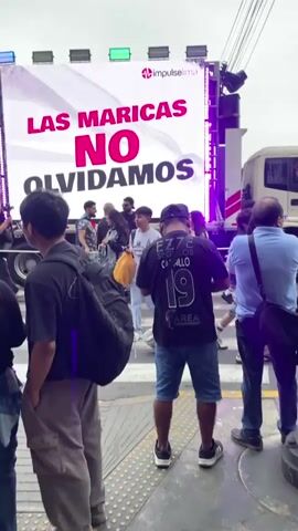 The video shows a large outdoor LED screen mounted on a truck. The screen displays pink text that reads "LAS MARICAS NO OLVIDAMOS" and then changes to "CREES QUE NO TE AFECTAN... HASTA QUE TE TOCAN A TI". There are several people in front of the truck, some of whom are looking at the screen. The environment appears to be an urban street, with overhead wires visible. Some people are holding what appear to be banners or flags. The overall mood seems to be one of a public event or demonstration.