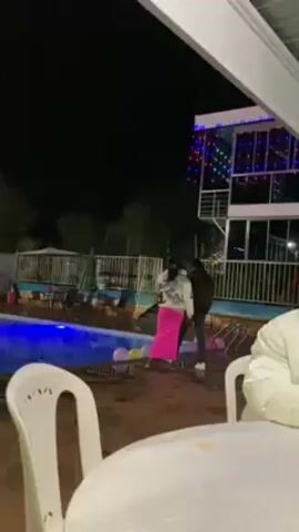 The video shows an outdoor swimming pool at night. A spotlight illuminates the pool area, and multicolored lights decorate a building in the background. The pool is surrounded by a tile patio with white plastic chairs and tables. The building next to the pool has a white observation deck and a mural. The camera pans from left to right, showing the pool and the surrounding area. Music plays in the background. The camera briefly focuses on a person wearing a white baseball cap before returning to the pool area.