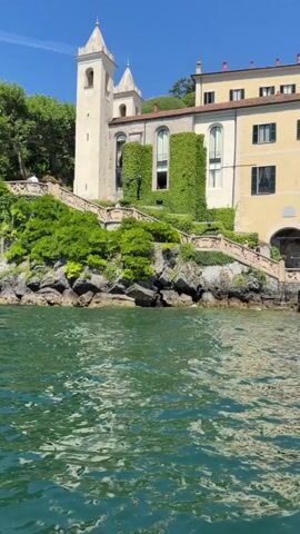 A drone shot captures a yellow and cream-colored building with green ivy climbing its walls, situated on a rocky incline overlooking a body of water. The building has a bell tower and several windows. Steps lead up from the water to the building, and lush green trees and bushes surround the property. The water in the foreground is a vibrant green with white ripples. The sky above is clear and blue. The shot pans slightly to the right, revealing more of the building and its surroundings.