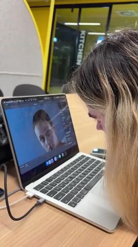 A woman with shoulder-length blonde and dark brown hair is sitting at a wooden desk, intently looking at a laptop screen. The laptop, which is connected to a charging cable, displays a video of a woman underwater. The environment appears to be a modern, brightly lit workspace with a partial view of a "KITCHEN" sign visible through a glass door in the background.