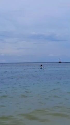 The video shows a lone person paddleboarding on a calm blue ocean. They are in the middle of the frame, relatively far from the camera. In the distance, there's a short lighthouse-like structure on a strip of land. The sky is overcast with a mix of light and dark blue hues. The overall scene is serene and minimalistic, highlighting the individual's solitude and connection with the water.