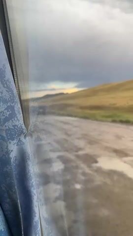 The video shows a view from a window, possibly of a vehicle, looking out onto a dirt road. The road is wet and has several puddles of water on it, indicating recent rain. The landscape is rolling hills covered in dry, golden-brown grass. The sky is cloudy with patches of bright light, suggesting a break in the clouds or the sun trying to emerge. A curtain, light blue with a white patterned design, is visible on the left side of the frame, obscuring part of the view. The camera appears to be moving, as the landscape shifts slightly throughout the short clip.