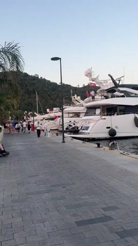This media file captures a lively scene at a marina in Marmaris, Turkey. The setting is a waterfront promenade paved with grey, interlocking bricks. On the right, a modern, white yacht is docked, its sleek lines contrasting with the more classic design of another yacht further down the pier. The yacht prominently displays the name "Melissim" in elegant script.

Several people are visible. On the "Melissim," two individuals stand on the upper deck, one waving with a raised arm, suggesting a celebratory or farewell gesture. Below them, a woman in a black dress is handing a yellow rope to a man in a blue t-shirt, indicating preparations for docking or departure. On the left side of the frame, a group of people stroll along the promenade, some appearing to be tourists. The time of day appears to be late afternoon or early evening, with the sky a soft, clear blue, hinting at pleasant weather. Lush green hills, characteristic of the Marmaris region, form a scenic backdrop.

Two utility boxes are present on the dock. One is blue with a yellow triangle and a symbol indicating a potential hazard. The other, to the right, is grey and also features a yellow warning sign with text in what appears to be Turkish, likely relating to electrical safety or a similar caution. The overall atmosphere seems relaxed and vibrant, typical of a popular tourist destination like Marmaris.