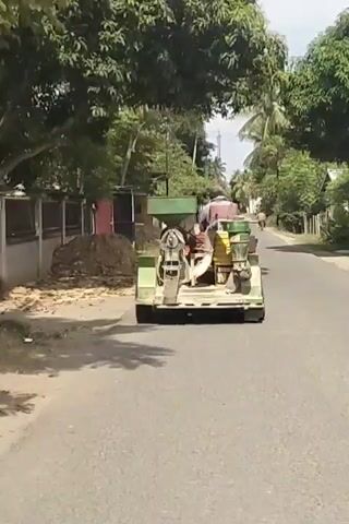 A motorized cart with a person driving it and a green hopper on the front is moving down a paved road. The cart has a metal frame and appears to be carrying some equipment. There are trees and houses on either side of the road. Another person on a motorcycle is visible in the distance behind the cart. The scene is outdoors during the daytime.