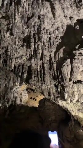 The video shows a dark cave illuminated by artificial light, revealing numerous stalactites hanging from the ceiling and stalagmites rising from the floor. Some formations are massive, appearing as columns. The camera moves through the cave, panning across the various rock structures, some of which are brightly lit, highlighting their texture and shape. Several people are visible in the lower part of the frame, their heads and shoulders silhouetted against the illuminated cave walls, suggesting they are on a guided tour or exploring the cave. One person is holding up a smartphone, likely recording or taking photos. The overall impression is one of awe and wonder at the natural formations.