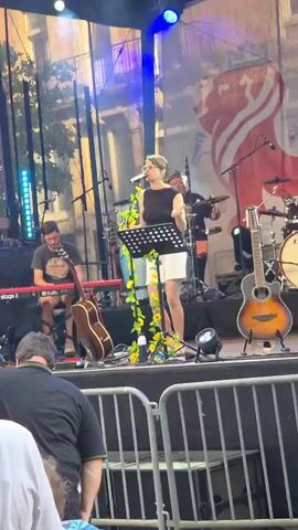 A group of musicians is performing on an outdoor stage at what appears to be a festival. In the foreground, a woman with short, dark hair is holding a young child wearing a red dress. The child is facing away from the camera. Behind them, a metal barricade separates the audience from the stage. The stage is set up with various musical instruments, including drums, guitars, and a keyboard. A female singer, wearing a black sleeveless top and a white skirt, stands center stage with a microphone and a music stand decorated with yellow flowers. A guitarist and a bass player are also visible on stage. The backdrop behind the musicians features a large banner with red and white graphics. The lighting suggests it is daytime or early evening.