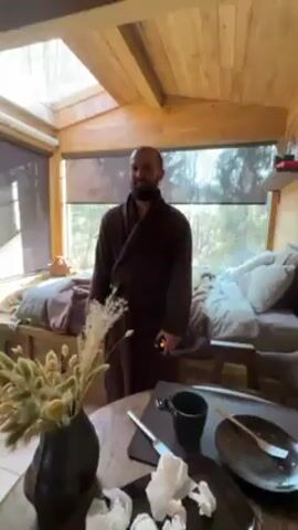 The video shows the interior of a small, rustic-looking cabin.  A man in a brown robe repeatedly steps onto and off a bed, with a determined look on his face. The bed, which seems to double as a seating area, is located near a large window that provides a view of trees. The room is brightly lit, with sunlight streaming in through the window and a skylight. In the foreground is a wooden table with a black vase containing some dried flowers, as well as dirty plates, cutlery, and a mug, suggesting a recent meal. The cabin's walls and ceiling are constructed of light-colored wood, giving the space a warm and cozy feel.