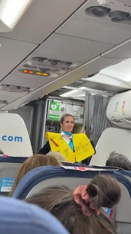 The video shows a flight attendant inside an airplane cabin, standing in the aisle between rows of seats. She is wearing a dark blue uniform with a light blue scarf and a bright yellow vest. The vest has writing on it in yellow marker. She holds up a yellow object, possibly a life vest or a demonstration prop, with writing on it, and gestures with her hands. The overhead compartments and lighting fixtures are visible above her. The heads of passengers are visible in the foreground, in the lower part of the frame. The background shows more of the cabin, including a closed curtained doorway.