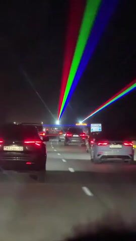 The video shows a nighttime view from inside a vehicle traveling on a highway. Multiple cars are visible on the road ahead, all appearing to be moving in the same direction. Above the highway, several bright laser beams of various colors, including red, green, and blue, are projected into the dark sky, creating a striking visual effect. The lasers appear to be emanating from a source that is not visible in the frame. The road is well-lit by the headlights of the cars and some overhead streetlights or signs. The overall impression is a dynamic and visually interesting scene, likely created for entertainment or as part of an event.