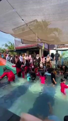 The video shows a large group of people, mostly children, gathered in and around a swimming pool, and the pool is partially shaded by a white canopy. Many of the children are in the water, with some wearing swimsuits and others in clothing. A small slide leads into the pool from one side, and some children are using it. Around the perimeter of the pool, a group of adults, many wearing hijabs, are standing and watching the children. The environment suggests an outdoor gathering, possibly a party or event centered around the pool. There appears to be a building with a red roof in the background, and some greenery visible beyond that.