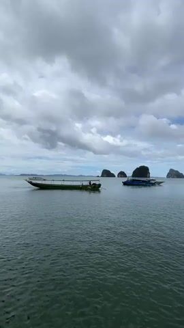 Two boats are on the water. One is a green boat on the left, and a blue and white boat is on the right. The green boat is long and narrow and has a canopy. There are people visible in the green boat. The blue and white boat is shorter and wider. There are islands in the background. The sky is cloudy. The water is choppy.