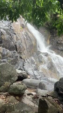 The video depicts a waterfall cascading down a rocky slope, framed by lush green foliage in the foreground. The waterfall itself is multi-tiered, with water flowing over gray and wet rocks. Large boulders are scattered at the base of the waterfall.  A small, square sign with an unknown logo is visible on the rock face above the lower level of the waterfall. The camera is stationary, providing a view from a lower angle looking upwards at the waterfall.