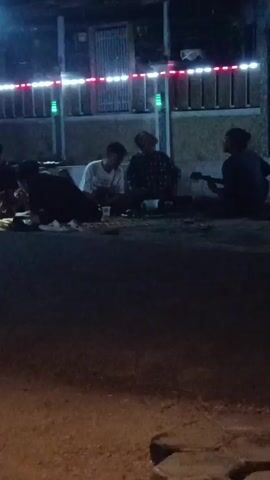 Four young men are seated on the ground outdoors at night, in front of a building. The building has a decorative string of lights along its upper edge, alternating between red and white, with some green lights interspersed. One of the men, wearing a dark shirt, is playing a guitar. The other three men appear to be listening or conversing. There are some cups and possibly food containers scattered on the ground in front of them. The ground is dirt or gravel, and a paved area is visible in the foreground. The lighting is dim, with the string lights providing the primary illumination.