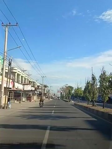 The video shows a view from a vehicle traveling down a street. On the left side of the street are buildings with shops. One shop sign is partially visible. There are power lines running overhead. A person is riding a motorcycle on the left side of the road. On the right side of the road are trees and more buildings in the distance. The sky is blue with some wispy clouds. The road appears to be paved with white lane markings. The overall impression is of a commercial street in a sunny climate.