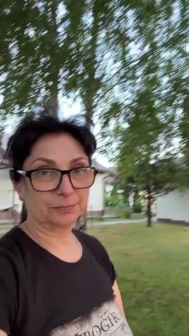 A short, shaky video displays a woman with short dark hair and glasses, wearing a black t-shirt with a graphic design. She is outdoors, with trees and grass visible. The background also shows white buildings. The woman looks directly at the camera and smiles. The video is filmed from a first-person perspective, indicating the woman is holding the camera.