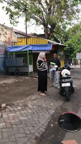 The video shows a street scene with two people standing near a food stall. A woman wearing a hijab and a black and white striped top is standing on the left. A man in a blue shirt and grey pants is on the right, holding a clear plastic bag. A motorcycle is parked next to the man. In the background, there is a yellow building with a blue awning covering the food stall. The ground is paved with bricks in the foreground and dirt or asphalt in the background. Trees are visible above the buildings. The overall lighting suggests it is daytime, possibly overcast.