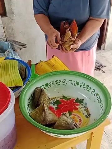 A person is shown preparing food. The person is holding a triangular-shaped item wrapped in banana leaves. In the foreground, a large bowl contains several similar items also wrapped in banana leaves. To the left, there are more banana leaves in a blue bowl and a plastic container with a red lid. The background appears to be an indoor setting with a textured wall.