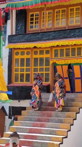 The video shows two people in traditional colorful costumes and masks dancing on a set of stairs in front of a building with yellow windows. The building is decorated with colorful banners. One of the dancers is holding a white object. The masks appear to be animal-like. The dancers are moving their arms and bodies in a synchronized manner. There are other people visible in the background, some of whom are also wearing white clothing and masks. The environment appears to be outdoors in a sunny location.