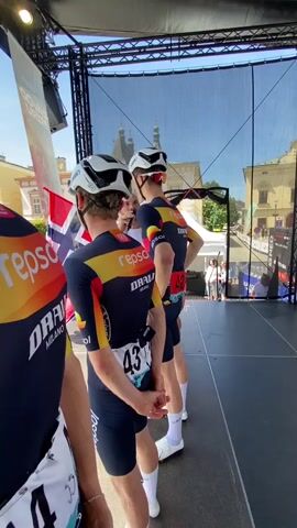 Cyclists stand on a stage being interviewed, wearing cycling gear with a number on the back. Spectators are in the square outside. A man is holding a microphone in front of the cyclists. They are standing in front of a black screen. The cyclists have white helmets on. The man interviewing them is wearing a white shirt. The cyclists have Repsol on their jerseys. The square is cobblestone. There are several white tents in the square. The sky is clear and blue. There are buildings on the perimeter of the square. A barrier separates the square from the stage. The cyclists are facing away from the camera.