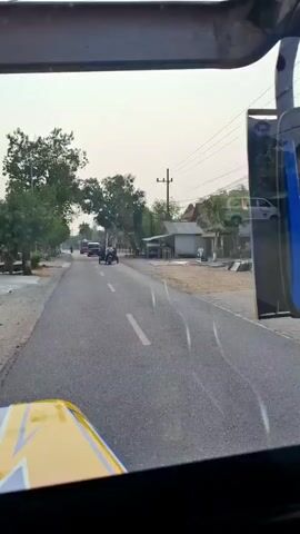 The video shows a view from inside a vehicle, looking out at a two-lane road. Several vehicles are visible on the road. In the foreground of the video, the hood of the vehicle the camera is in is visible. It is yellow and black. The road is asphalt and has a white dashed line down the center. On the left side of the road, there are trees and some buildings. On the right side of the road, there are more buildings and power lines.

In the distance, a motorcycle with a sidecar is carrying a person and a long, thin object, possibly a pole. Behind it, another motorcycle is visible. Further down the road, a dark gray van is driving towards the camera. To the left of the van, a smaller truck is also driving towards the camera.

The scene appears to be a rural or suburban area, possibly in Southeast Asia, given the types of vehicles and the style of buildings. The sky is overcast. The overall impression is of a calm, ordinary day with local traffic.