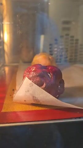 The video shows the baking process of several dough items inside an oven. Initially, a red-colored dough ball, possibly cookie dough, is prominently displayed in the foreground, resting on parchment paper and a red silicone mat. An out-of-focus, raw, orange-colored dough ball is visible in the background. As the video progresses and the camera pans slightly, another orange dough ball comes into view. These orange dough balls gradually puff up and transform into a golden-brown, baked state. The red dough ball also appears to expand slightly during the baking but maintains its red color. The interior of the oven, with its metal walls and a visible light source on the left, provides the setting for this transformation. There is no visible text.