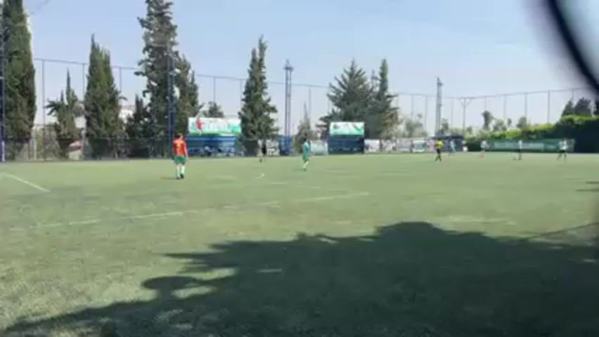 The video shows a soccer match on an outdoor artificial turf field on a sunny day. Several players wearing different colored uniforms are spread across the field. On the left is a player in an orange and green uniform. Several players in light green uniforms are positioned near the center of the field. A few players in dark uniforms are towards the right. A tall fence surrounds the field, with tall trees visible behind the fence. Advertising banners line the backdrop of the field. Shadows from the fence and trees are cast on the field. A partial view of a fence obstructs the camera's view in the foreground.