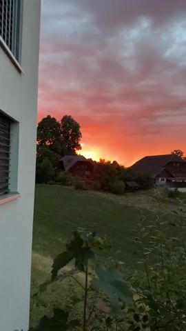 A video shows a scenic view of a rural landscape at sunset. The sky is ablaze with vibrant hues of orange, red, and pink, with dramatic clouds casting shadows over the scene. In the foreground, lush green grass covers a rolling hill. Several houses with traditional architecture are nestled amongst trees, creating a picturesque village atmosphere. A close-up shot of leaves in the foreground adds a touch of nature's beauty to the composition. The video captures the essence of a peaceful evening in the countryside, bathed in the warm glow of the setting sun.
