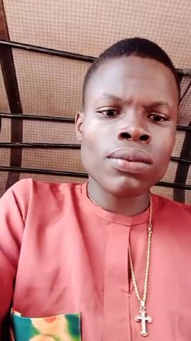 A close-up, eye-level video shows a young man in a reddish-orange, button-up shirt with a gold chain and cross pendant. He looks directly at the camera with a serious expression. Behind him, the patterned ceiling of a vehicle, possibly a tuk-tuk or auto-rickshaw, is visible. For a brief moment, a second person is seen to the left, appearing to yawn. The young man remains the primary focus throughout the video.