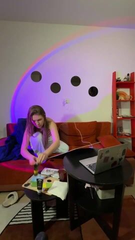 A young woman is sitting cross-legged on a red-orange couch, squeezing black paint from a tube onto a palette on a small black table. She's wearing light-colored pajamas. Next to the palette is a towel. A laptop sits open on another small black table. Behind the woman are four round mirrors on a white wall, illuminated by a rainbow light effect. To the right is a red bookshelf. The room is lit by the rainbow light and overhead lighting. A blanket and slippers are visible on the couch.