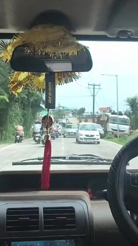 The video is shot from the interior of a car, looking forward through the windshield. In the foreground, the dashboard and steering wheel are visible. Hanging from the rearview mirror are two decorative items: a gold garland with spiky protrusions and a black air freshener with a red tassel. The road ahead is congested with traffic. Several motorcycles and cars are moving slowly. On the left side of the road, there are trees and lush green foliage. On the right side, buildings and utility poles line the road. The sky is overcast, suggesting a cloudy or potentially rainy day. There are people on the motorcycles, some wearing helmets. The overall impression is a busy street scene in a moderately populated area, possibly a town or city in a hilly or mountainous region, given the vegetation.
