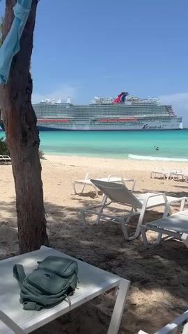The video opens with a wide shot of a beach, showing lounge chairs in the foreground. A large cruise ship is visible in the turquoise water, with a few people swimming near the shore. The camera pans to the right, revealing more lounge chairs and trees providing shade. Another shot shows a person relaxing on a lounge chair on the beach, facing away from the camera. The scene is idyllic, with clear blue skies and calm waters. The camera continues to pan, showing a lifeguard stand with a red umbrella and more people enjoying the beach. The final shot is a close-up of a person lying on a lounge chair, appearing to be asleep, with the beach and ocean in the background.