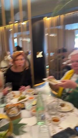The video shows a group of women having lunch at a restaurant. They are seated at a table covered with a white tablecloth patterned with a large "K" design. The table is laden with various dishes of food, including sushi, dumplings, and other Asian-inspired cuisine. There are also several glasses, bottles of water, and decorative elements on the table.

The women are dressed in casual attire. They are interacting with each other, reaching for food, and seemingly engaged in conversation. The camera moves around the table, showing different angles of the scene and capturing the faces of the women. The restaurant has a modern decor with wooden panels and ambient lighting. The overall atmosphere appears to be relaxed and social.