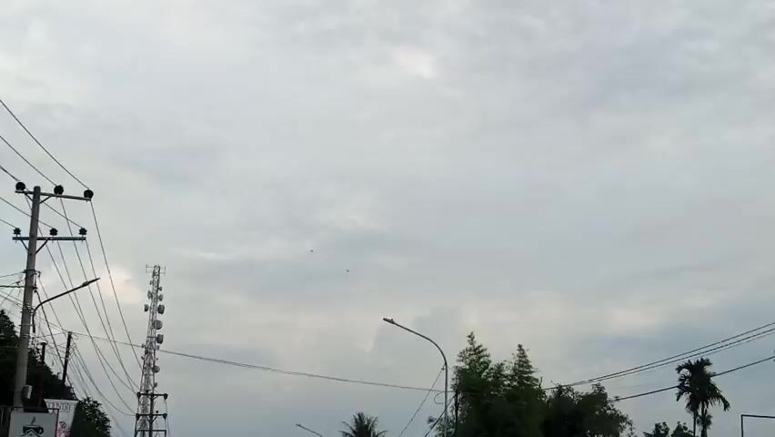 The video captures an overcast sky with a cluster of clouds. In the foreground, a tall cell tower is visible on the left side, with multiple antennas attached to it. A utility pole with electrical wires extends across the frame. To the right of the cell tower, a street light is mounted on a curved pole. Behind the street light, a dense cluster of dark green trees obscures the view. Further to the right, parts of two roofs are visible, suggesting the presence of buildings. The overall lighting is dim, consistent with a cloudy day. The camera appears to be moving slightly, creating a subtle panning effect.