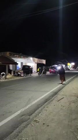 The video shows a dark, outdoor scene at night. A paved road runs through the center of the frame, with a shoulder on the right side. A woman stands on the shoulder, facing the road, with her back to the camera. She is wearing a dark hijab and a patterned skirt. Her shadow is cast on the road. On the left side of the road, there are buildings that appear to be shops or businesses, some with illuminated signs. Several vehicles are visible on or near the road, including a pickup truck, two motorcycles, and a pink car. One motorcycle is in motion, heading away from the camera. Another motorcycle appears to be parked. A cart or food stall with a blue and yellow canopy is visible on the far left. The overall environment suggests a roadside commercial area at night.