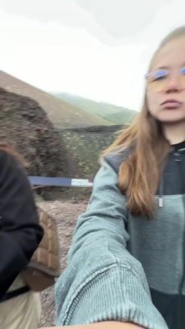 The video shows a young woman on a rocky, volcanic landscape, possibly a mountainside. The environment is barren with dark, red and black rocks. She is wearing glasses, a gray and black jacket, and has long brown hair. She holds the camera at arm's length, taking a selfie. Initially, she looks directly at the camera with a serious expression. She then makes a peace sign and sticks out her tongue. In the background, other people are walking up a steep incline of the volcanic terrain. At one point, a man in a blue jacket and baseball cap walks past her. The sky is overcast and misty. The camera view is generally shaky, indicating the person is moving slightly.