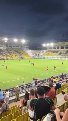 This video captures a moment from a football match, likely played at the Sheriff Stadium in Tiraspol, Moldova. The scene is set in the evening, with the stadium lights illuminating the lush green field. The sky above is a deep, dusky blue, suggesting the game is either at dusk or under floodlights.

The focus is on a section of the crowd seated in bright yellow tiered seating, looking towards the pitch. Spectators of various ages are visible, some standing and watching the game, others seated and engaged. Their attire is casual, typical for a sporting event. The camera angle gives a perspective from behind the spectators, looking over their shoulders and heads towards the action on the field.

On the field, players in red and black kits are engaged in a game. The stadium itself appears to be a modern facility with white and yellow accents on its structure, and a large capacity crowd can be seen in the stands across the field. The overall atmosphere seems to be one of lively engagement, with people actively watching the football match unfold.

There is no visible text in this video, but the context provided by the prompt places the event in Tiraspol, Moldova.