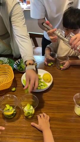 The video shows a group of people, including at least one child, gathered around a wooden table preparing drinks. In the foreground, several clear plastic cups are filled with sliced green fruit, likely limes or lemons, and possibly other ingredients. A yellow bowl contains more sliced green fruit and a whole yellow lemon. To the left, a yellow basket holds more green fruits, and a plate displays whole cucumbers and lemons. A person in a white t-shirt is holding a metal device, possibly a muddler or juicer, and is pouring liquid into two cups in front of a child wearing a light-colored shirt. Another person in a light-colored jacket is adding slices of green fruit to a cup. The scene appears to be an informal setting, perhaps a kitchen or dining area, with a focus on making fresh beverages. The lighting is natural and highlights the vibrant green of the fruit.