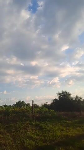 The video shows a cloudy sky with patches of blue. In the foreground, there is tall green grass and a wire fence. A wooden pole with a concrete base stands vertically in the middle of the frame, with a wire attached to it, extending out of frame. Further in the background, there is a brick house with a dark roof, surrounded by trees. The sun is setting, casting a warm glow on the scene.