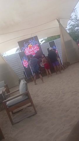 The video depicts a DJ booth set up on a sandy beach under a tent-like structure, with a sign behind it that says "Waikiki Semana Santa 2026". Several people are standing around the booth. A woman in a red bikini top and wrap skirt is facing the booth, while a man in a black t-shirt and purple shorts stands beside her. A man in a baseball cap playfully taps the woman's shoulder. Other people stand by, including one taking photos with a phone. The overall scene has a relaxed, beach party atmosphere.