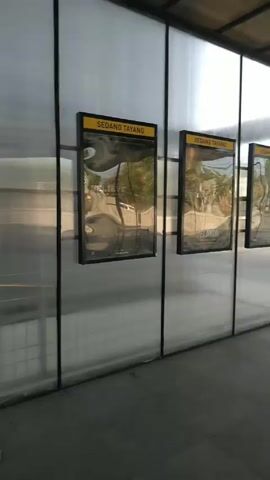 The video shows a series of movie posters displayed along a walkway. There is a poster for a film called "La Tahzan: Cinta, Dosa, Luka..." advertising its release on August 14, 2025. Alongside it are framed posters labeled "Sedang Tayang" and "Segera Tayang," though their titles aren't fully visible. One of the "Segera Tayang" posters appears to be for a movie called "Lyora." A wooden and metal bench sits in front of the posters. The walkway's walls are made of semi-transparent white panels with a black frame.