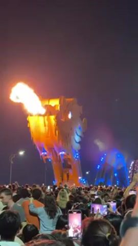 A large, illuminated dragon statue emits a large plume of fire from its mouth in this outdoor nighttime scene. The dragon is tan-colored with blue lights outlining its scales. It stands on a bridge over water. A large crowd is gathered in front of the dragon, many holding up their phones to record the display. The sky is dark, and streetlights line the background. After emitting fire, the statue begins to spew smoke.